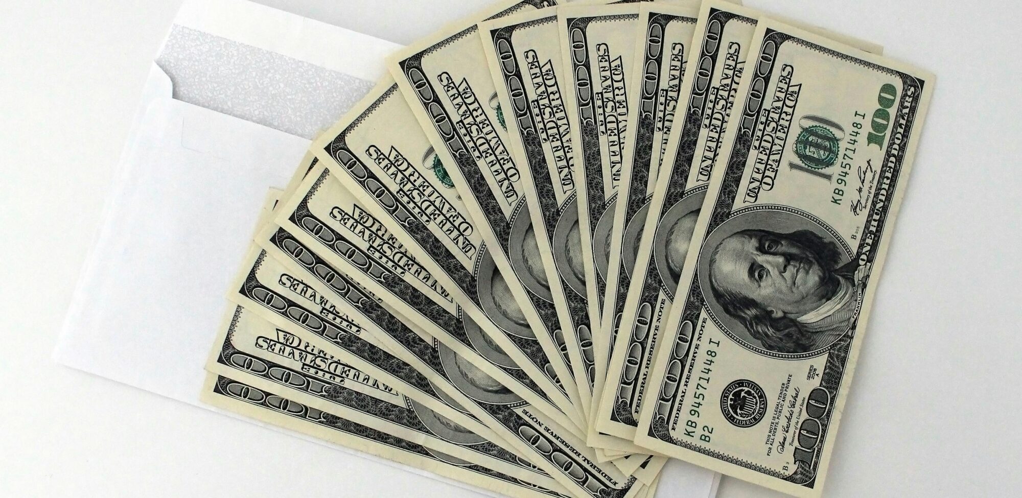 Fan of US $100 bills partially out of a white envelope on a white background.